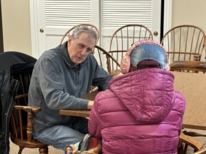 A man in a blue sweater helps a woman in a pink coat fill out a questionnaire as they are seated at a table