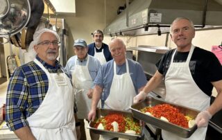 Five men in a kitchen wearing aprons, with some holding trays of food