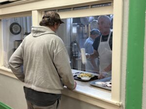 A man in a gray sweatshirt and hat stands at a counter with a glass wall, behind which a man in an apron serves him food