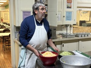 A man wearing a blue shirt and white apron standing over bowls of food in a shelter kitchen