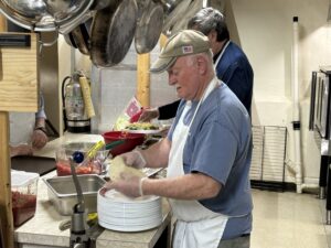 A man wearing a blue shirt and white apron prepares food while standing next to another man in a kitchen